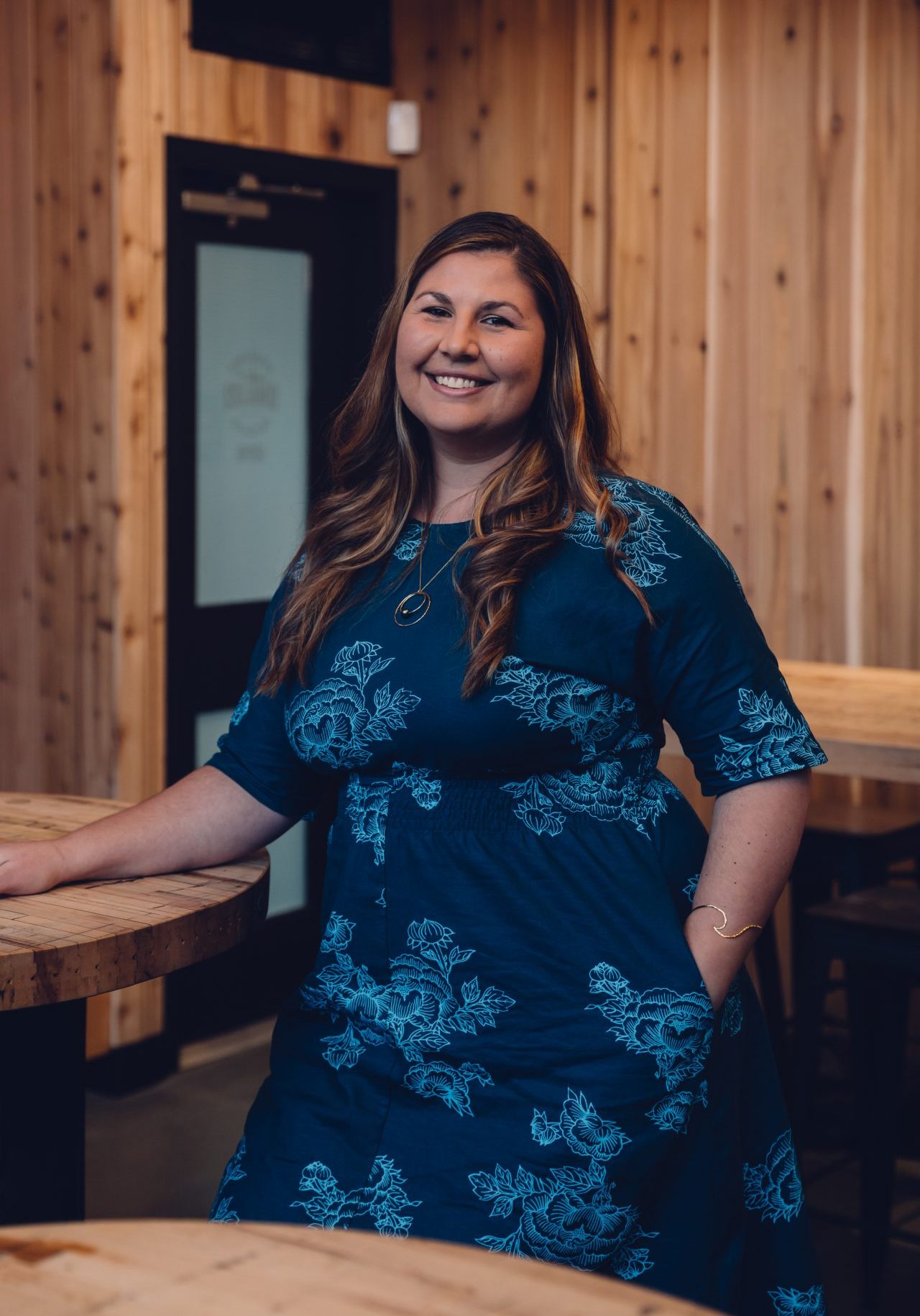 Operations Manager & Training Facilitator Tierra Lavery is a woman with long, brown hair with highlights. She is wearing a blue dress with a floral design, and smiling warmly into the camera.